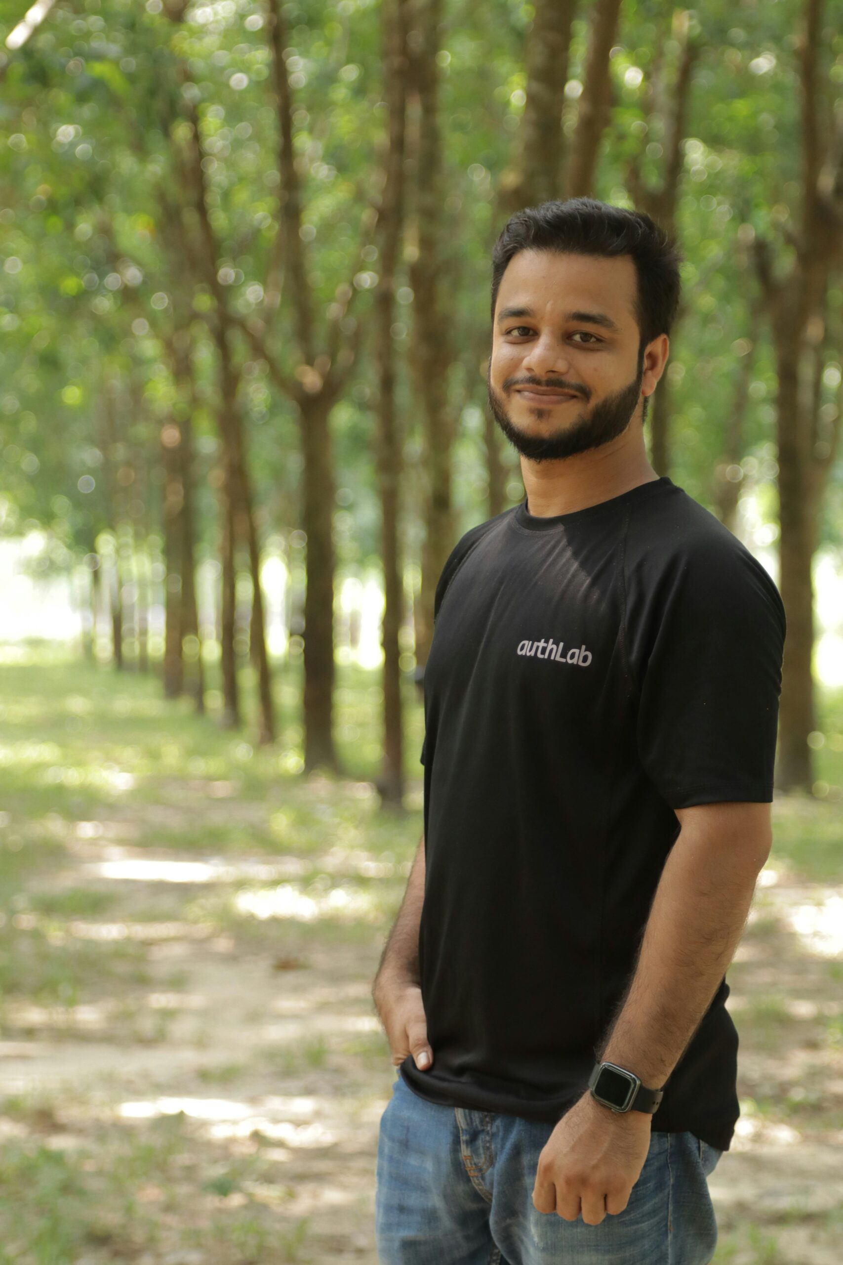 A happy man wearing a black shirt smiles in a tree-lined park setting.
